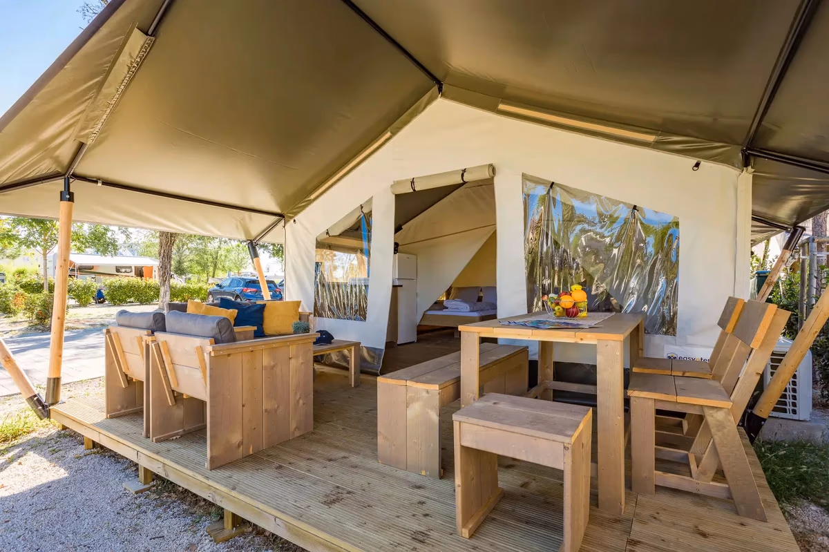 Overdekt terras van een Family Glamping Tent op Camping Lanterna met houten loungebank, tafel met krukjes en doorkijk naar het bed