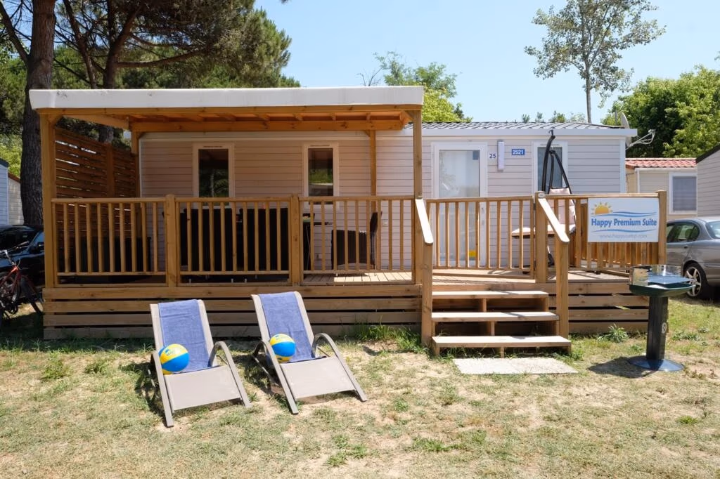 Happy Premium Suite op Camping Lanterna met een groot houten terras, naambordje en twee strandstoelen met handdoeken op het gras