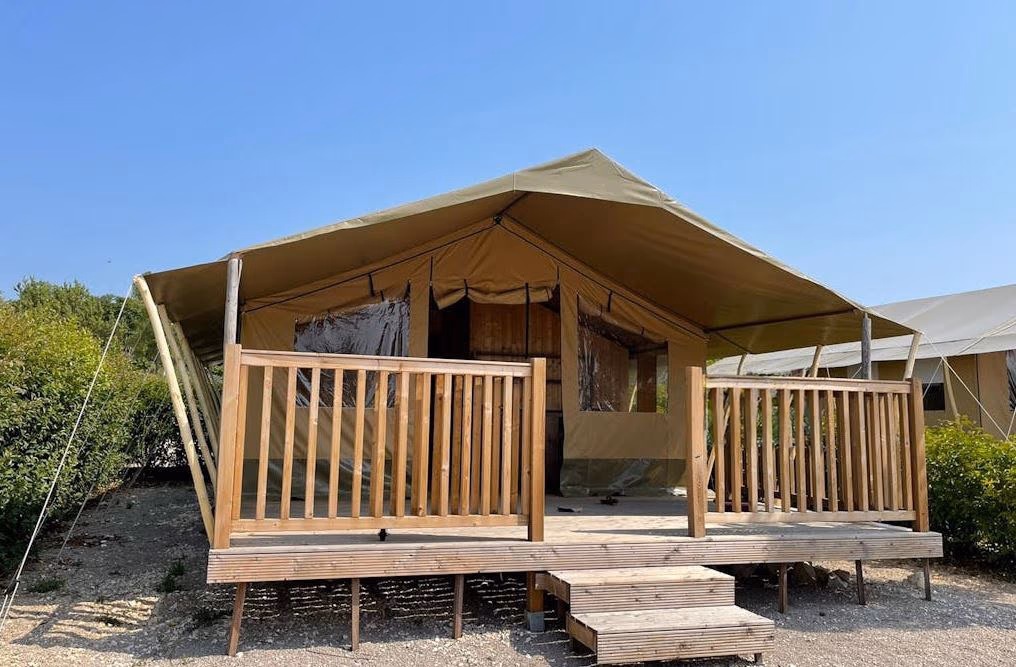 Safaritent Sorella op Camping Lanterna met een beige dak en een houten veranda met reling en trap, tegen een strakblauwe lucht