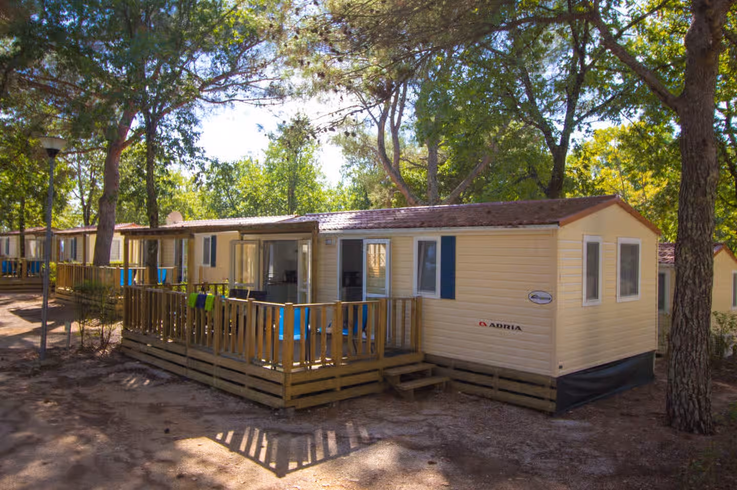 Gele stacaravan Malta met blauwe luiken en een houten vlonderterras, tussen de dennen op Camping Lanterna