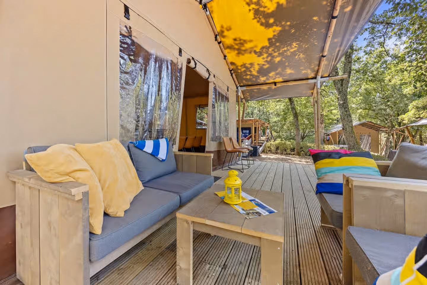 Lang overdekt houten terras van een grote lodgetent, met grijze loungebanken en gele en gestreepte kussens