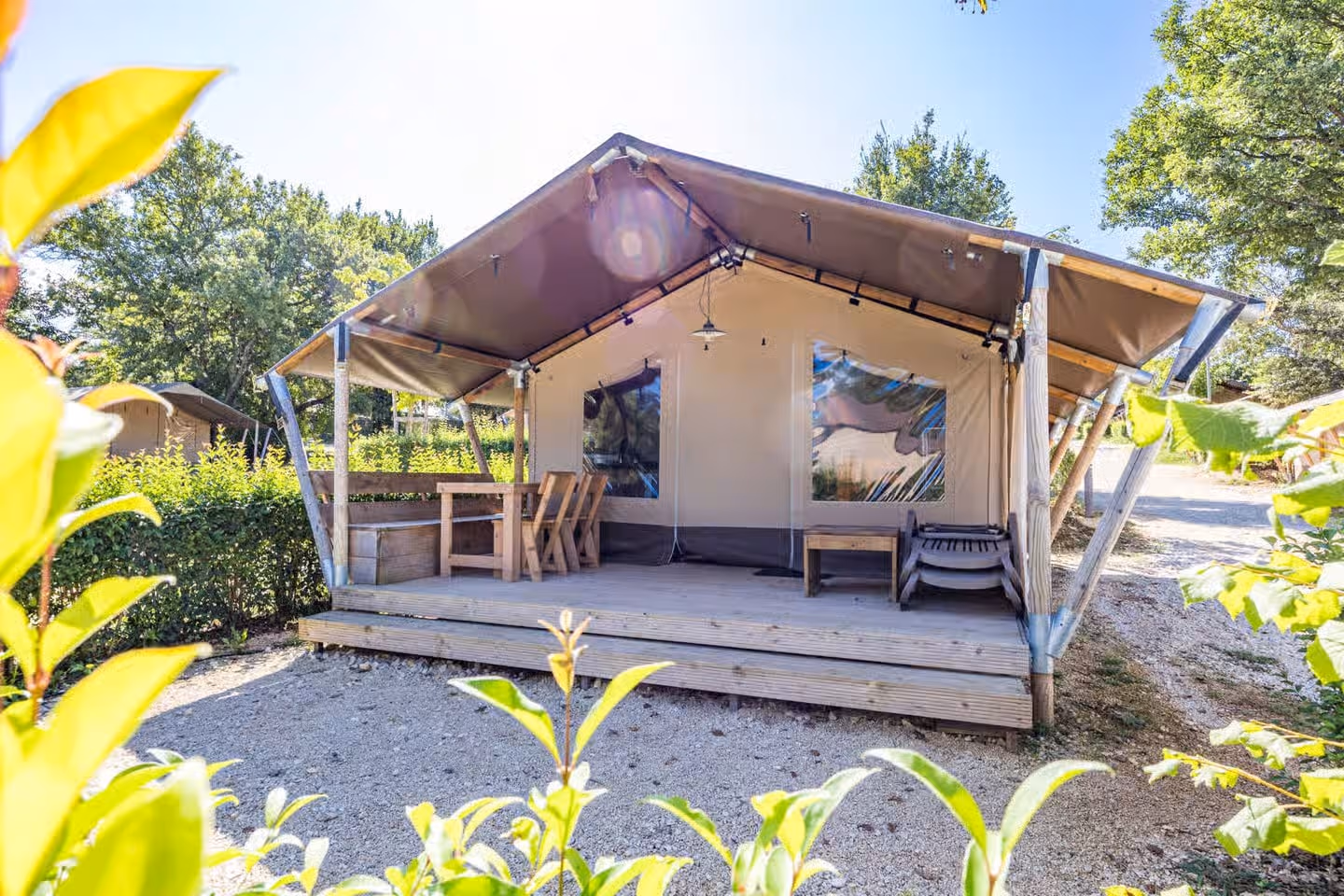 Safaritent met een overdekt houten terras met eettafel en stoelen, op een grindplek met struiken