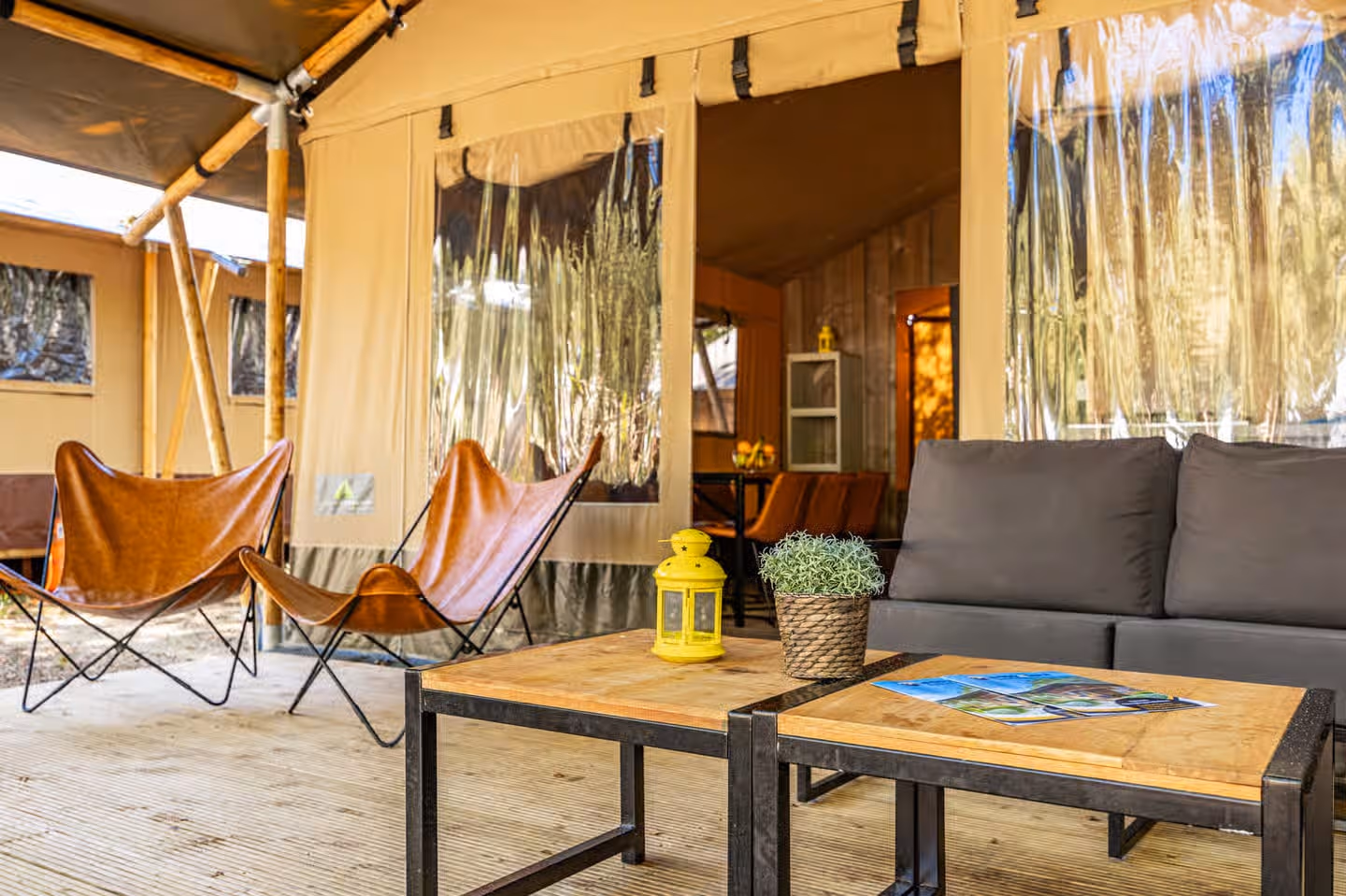 Overdekt terras van een lodgetent met twee leren vlinderstoelen, een grijze loungebank en houten salontafels
