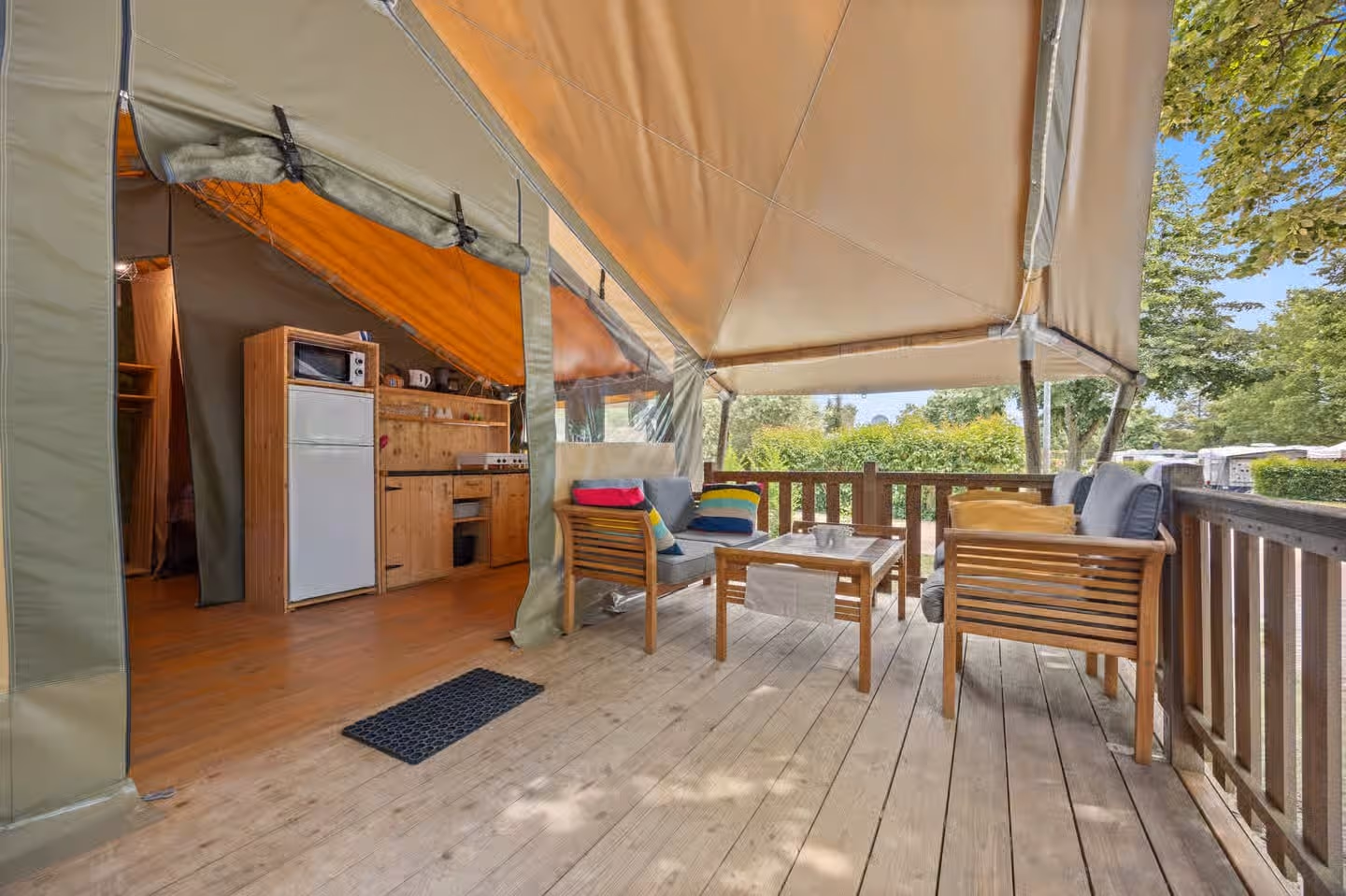 Overdekt houten terras van een Safaritent Relax, met tuinstoelen en een salontafel en de keuken met koelkast in de tent erachter