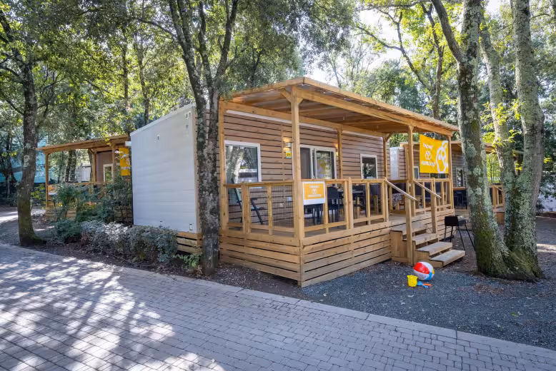 Premium-stacaravan met houten gevel en overdekt terras onder hoge eiken op Camping Lanterna, met een gele Eurocamp-vlag