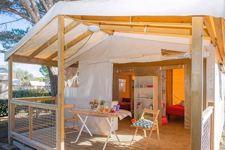 Safaritent met een houten luifel en een klaptafel met bloemen op het terras, en een doorkijk naar de open tent met een rek en bedden