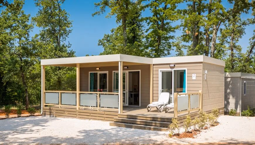 Taupekleurige Premium-stacaravan met plat dak, overdekt terras en twee ligbedden, op een grindplek tussen de eiken op Camping Lanterna