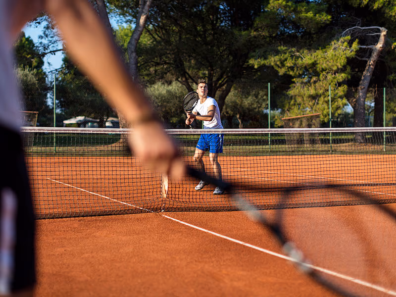 Tennissers op de gravelbaan tussen de pijnbomen bij het sportcentrum van Camping Lanterna