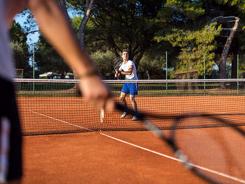 Tennissers op de gravelbaan tussen de pijnbomen bij het sportcentrum van Camping Lanterna