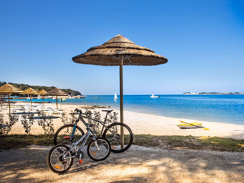 Twee fietsen onder een rieten parasol aan het strand Sunce van Camping Lanterna