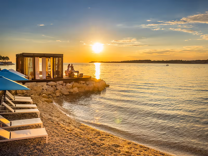 Zonsondergang bij het Marbello-strand van Camping Lanterna, met houten strandcabana en blauwe ligbedden